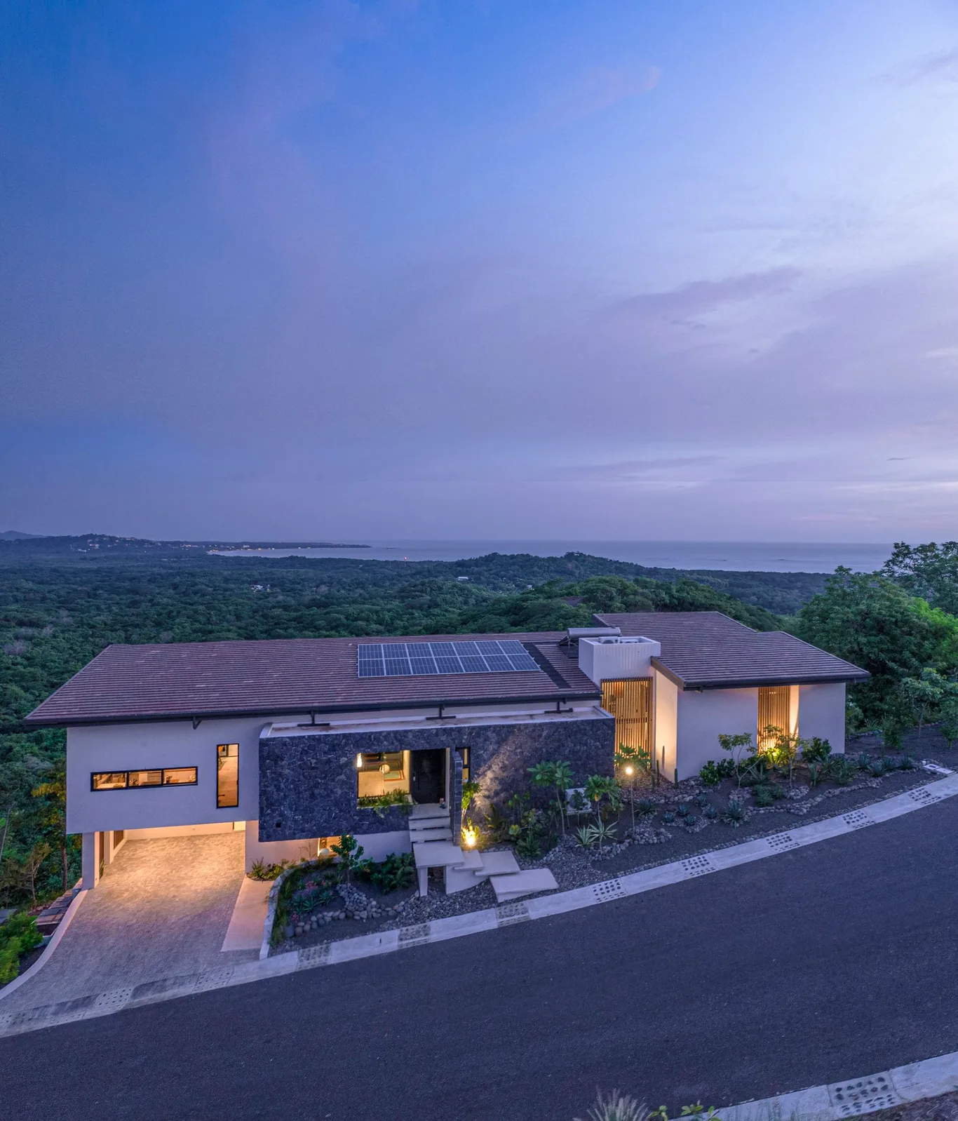 Home at dusk with a stone entrance, solar roof and dry garden, the forest and the Pacific coast beyond